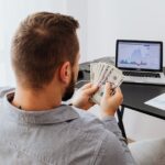 A man is sitting infront of a laptop with a currency notes in his hands.