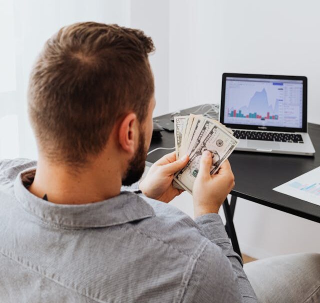 A man is sitting infront of a laptop with a currency notes in his hands.
